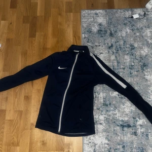 Mörkblå DRI FIT från Nike - Säljer en mörkblå Nike Dri-Fit träningsjacka med vit dragkedja och vit rand längs ena ärmen. Jackan har lång ärm och är tillverkad i ett lätt syntetmaterial som andas. Perfekt för träning eller sportiga aktiviteter.