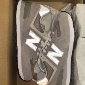 Newbalance Classic 574 - Säljer ett par klassiska grå New Balance sneakers med vit sula och stora vita N-loggor på sidorna. Skorna har snörning och är tillverkade i mocka och mesh för en sportig och stilren look. Perfekta för dig som gillar streetstyle.