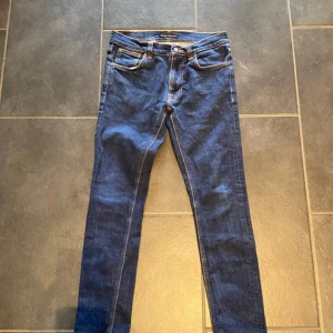 Blå slim fit jeans från nudie  - Snygga blå skinny jeans från nudi  med klassisk femficksdesign och kontrastsömmar. Jeansen har normal midja och smal passform hela vägen ner. Bakfickorna har en gul broderad detalj som sticker ut. Perfekta till sneakers eller boots.