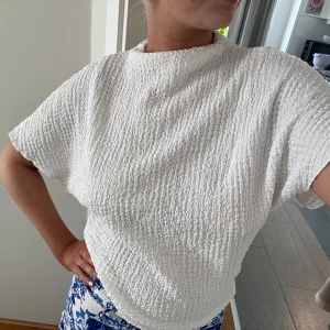 Vit strukturerad blus  - Säljer en vit blus med kort ärm och hög hals. Toppen har en snygg strukturerad, nästan krinklad yta som ger en cool och modern känsla. Passformen är lite loose och materialet känns mjukt och luftigt, perfekt för varmare dagar.