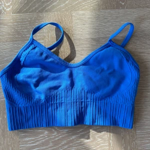 Blå ribbad linnetopp med smala band - Snygg blå linnetopp med smala axelband och ribbade detaljer nedtill. Toppen är ärmlös och har en tight passform som sitter skönt mot kroppen. Perfekt för träning eller som ett coolt basplagg till vardags.