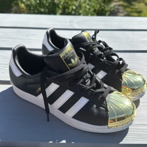 Adidas Superstar med guldtå - Svarta och vita Adidas Superstar sneakers med ikoniska tre ränder och metallic guldfärgad tå. Skorna har snörning, platt sula och detaljer i guld på tungan och sidan. Tillverkade i skinn och syntet, perfekt för dig som vill sticka ut med en unik twist på klassikern.