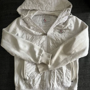 Vit quiltad jacka från Moncler - Snygg vit jacka från Moncler med quiltade partier framtill, huva och dragkedja. Jackan har ribbade muddar, två fickor med dragkedja och broderad logga på bröstet. Perfekt för dig som gillar stilren och sportig look.