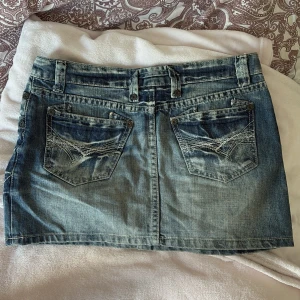 Blå lågmidjad jeanskjol med broderade fickor - Snygg blå lågmidjad jeanskjol i kort modell med slitna detaljer och broderade bakfickor. Klassisk femficksdesign, bälteshällor och knappstängning framtill. Perfekt för dig som gillar en avslappnad och cool stil. 