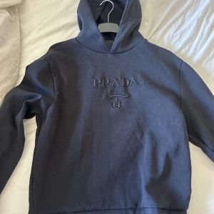 Svart hoodie från Prada - Snygg svart hoodie från Prada med broderad logga på bröstet. Klassisk modell med huva och långa ärmar.