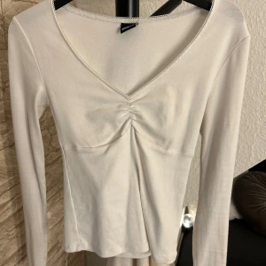 Vit långärmad topp från Gina Tricot - Snygg vit långärmad topp från Gina Tricot med v-ringning och rynkdetalj framtill. Mjuk och stretchig bomullskänsla, perfekt till jeans eller kjol. 
