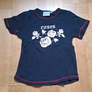 Svart t-shirt med rosor - Svart t-shirt från KappAhl med vita rosor och texten 'roses' framtill. Röda sömmar längs kanterna. Barnstorlek men passar xs, eventuellt s. 😊🩷