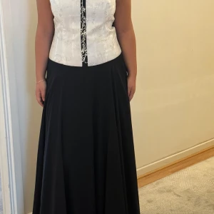 Svart och vit långklänning med broderi - Snygg långklänning med svart, vid kjol och vit, broderad överdel med tunna axelband. Överdelen har dekorativa detaljer och en smal svart kant framtill. Perfekt för bal eller student. Kjolen faller mjukt och ger en elegant siluett.