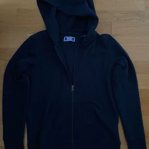 Svart hoodie från Jack & Jones - Svart hoodie från Jack & Jones med dragkedja hela vägen och huva. Tröjan har långa ärmar och praktiska fickor framtill. Perfekt för en chill och avslappnad stil. Tillverkad i mjuk bomull för extra komfort.