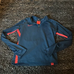 Nike Dri-Fit blå långärmad träningströja - Nike Dri-Fit långärmad träningströja i marinblått med orange- röda detaljer längs sidorna och på axlarna. Tröjan har half zip framtill och är tillverkad i ett lätt, syntetiskt material som andas. Perfekt för dig som gillar sportig stil och vill ha något bekvämt till träningen. Tröjan är i L men i barnstorlek! 147-158
