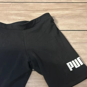 Svarta träningstights shorts från Puma - Svarta tighta träningstights shorts från Puma med vit logga på ena benet. De har en elastisk midja och är gjorda i ett stretchigt syntetmaterial som passar perfekt för sport och aktivitet. Snygga och enkla att matcha med din träningsoutfit.