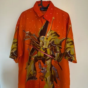 Orange Space Warriors skjorta med robotmotiv - Fet orange skjorta från Space Warriors med stort tryck av en gul robot på både fram- och baksidan. Skjortan har korta ärmar, klassisk krage och knäppning framtill. Perfekt för dig som gillar anime eller sci-fi vibes. Materialet känns som bomull och trycket är riktigt detaljerat.