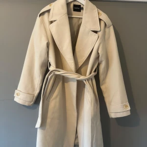 Beige kappa från Boohoo med bälte - Snygg beige kappa från Boohoo med breda slag, axelklaffar och knappar vid ärmslut. Kappan har bälte i midjan och två fickor framtill. Perfekt för dig som vill ha en stilren och trendig look under höst och vinter.