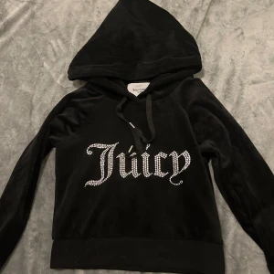 Svart velour hoodie Juicy Couture XS - Svart hoodie i mjuk velour från Juicy Couture med stor huva och snörning. Framsidan har glittrande stenar som bildar texten 'Juicy'. Långärmad och med klassisk relaxed passform, perfekt för dig som gillar en ikonisk Y2K-stil.