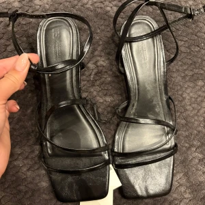 Svarta klackskor med smala remmar - Snygga svarta klackskor med smala remmar och fyrkantig tå. Skorna har låg klack och justerbar ankelrem. Tillverkade i syntetmaterial med extra komfort inläggssula. Perfekta för dig som gillar minimalistisk och trendig stil.