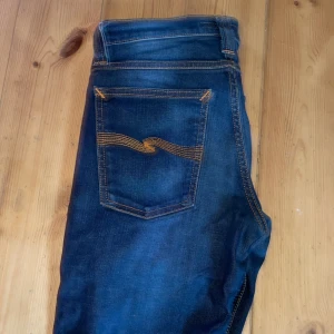 2 Feta Nudie Jeans blå - Säljer ett par klassiska blå jeans från Nudie Jeans med snygga kontrastsömmar och ikonisk broderad bakficka. Jeansen har läderpatch med logga och är tillverkade i bomull. Perfekta för dig som gillar stilrena och tidlösa jeans.