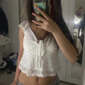 Vit spetsig croppad blus med knyt - Söt vit blus i spets med croppad passform och volangärmar. Toppen har en feminin v-ringning och knyt framtill som ger en romantisk vibe. Perfekt för varma dagar och lätta outfits.