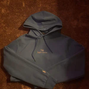 Blå hoodie från Polo Ralph Lauren - Snygg blå hoodie från Polo Ralph Lauren med broderad logga på bröstet och liten häst på ärmen. Tröjan har huva med snörning och är tillverkad i mjuk bomull. Perfekt för en chill och stilren look.