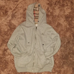 Grå Burberry hoodie med dragkedja - Snygg grå hoodie från Burberry med klassiskt rutigt foder i huvan. Storlek M men sitter mer som S. Den är i bra skick inga fläckar, märken, osv. Däremot kan dragkedjan strula lite ibland. Skriv om ni har frågor, hellre för många frågor än för få!😁