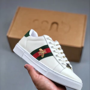 Gucci Ace vita sneakers med bi-detalj - Gucci Ace sneakers i vitt skinn med grönt och rött band på sidan, broderad guldfärgad bi och grönt krokopräglat läder på hälen. Klassisk rund tå, snörning och platt sula. Lyxig och ikonisk design med tydliga Gucci-detaljer.