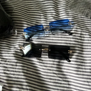 Två par rektangulära solglasögon - Säljer två par trendiga solglasögon med rektangulära glas. Ena paret har blåtonade linser och det andra har svarta linser. Båda har tunna guldfärgade detaljer och svarta skalmar. Perfekta för dig som vill sticka ut med en modern look.