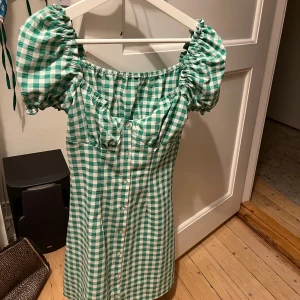 Grönvit rutig kortklänning med puffärm - Supersöt kortklänning i grönt och vitt rutigt mönster med puffiga korta ärmar och knappar framtill. Offshoulder-modell med markerad byst och lätt A-linjeformad kjol. Perfekt för sommarens alla tillfällen!