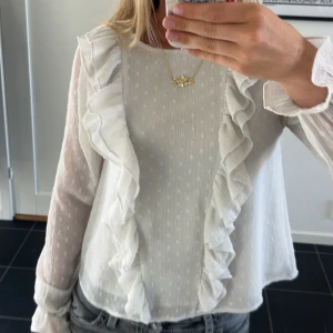 Vit blus med volangdetaljer - Supersöt vit blus med långa ärmar och stora volanger framtill. Tunt, lätt material med diskret prickstruktur och transparenta ärmar. Perfekt för dig som gillar romantisk och trendig stil.