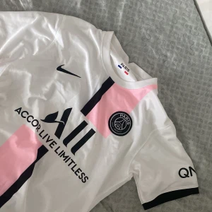 PSG vit och rosa Nike fotbollströja M - Snygg PSG fotbollströja från Nike i vitt med svarta och rosa detaljer. Tröjan har korta ärmar, klubbmärke på bröstet och Dri-FIT-material som andas. Paris Saint-Germain-logga och sponsortryck framtill. Perfekt för dig som älskar fotboll och vill sticka ut.pris kan diskuteras vid snabbaffär.