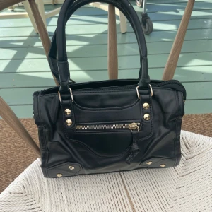 Svart handväska med nitar och dragkedja - Cool svart handväska i skinnimitation med snygga guldfärgade nitar och detaljer. Väskan har dubbla handtag, en dragkedja framtill och dekorativ tofs.  Ok skick med viss täcken på användning 