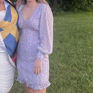 Ljuslila blommig klänning från Vila - Supersöt ljuslila kortklänning från Vila med småblommigt mönster. Klänningen har v-ringning, långa ärmar med volangdetalj vid ärmslut och volangkant nertill. Perfekt för sommarens alla tillfällen!