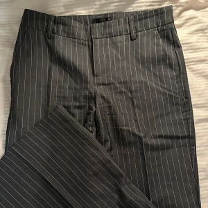 Randiga kostymbyxor från Gina Tricot - Snygga mörkgrå kostymbyxor från Gina Tricot i storlek 34. Byxorna har smala vita ränder. Perfekta för en classy outfit. Använt 1 gång