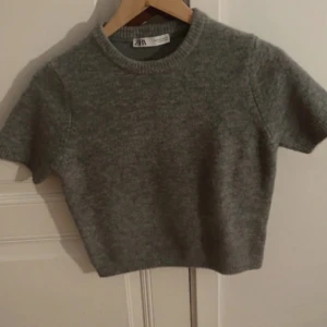Grå stickad croppad tröja från Zara - Grå stickad tröja från Zara med kort ärm och croppad passform. Tröjan har rund halsringning och är perfekt för lager-på-lager. Enkel och stilren design som funkar till många olika looks.
