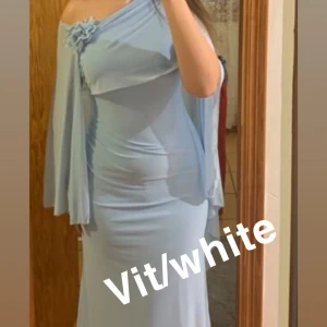 Vit långklänning med cape och blomdetalj - Säljer en vit långklänning med elegant cape-liknande överdel och en söt blomma på axeln. Klänningen har en flowy känsla och är figurnära med asymmetrisk fåll. för dig som vill sticka ut med en unik och stilren look. klänningen är använd en gång vid ett tillfälle så klänningen är jättefint skick. Man kan ha en långärmad tröja under ifall man inte vill visa armarna. Klänningen är nytvättad och är lite smutsig från när jag har gått längst ner på klänningen så ingenting man ser så noga kolla sista 