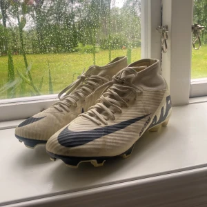 Nike Mercurial Air Zoom fotbollsskor beige - Säljer ett par Nike Mercurial Air Zoom fotbollsskor i beige med svarta detaljer och stor Swoosh-logga. Skorna har snörning, högre skaft och platt sula med dobbar för gräsplan. Lätta och smidiga i syntetmaterial, perfekta för dig som vill ha speed på planen. Storlek 41