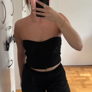 Svart ribbad bandeau tubtopp - Säljer en svart bandeau tubtopp med ribbad struktur. Toppen är axelbandslös och croppad, perfekt för dig som gillar en enkel men snygg stil. Sitter tight och har en stilren look som passar till många outfits. Mindre i storlek så passar även xs beroende på önskad passform. 