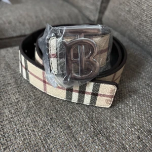 Burberry bälte - Snyggt bälte från Burberry med klassiskt rutigt mönster i beige, svart och rött. Bältet har ett stort metallspänne med ett B framtill. Perfekt accessoar för att lyfta din outfit med en lyxig touch.