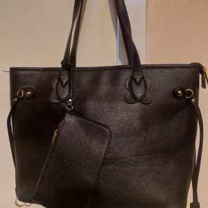 Svart toteväska från Chiquelle - Stilren svart axelväska i imitationsläder med guldiga detaljer och tillhörande clutch. Väskan har dubbla handtag, dragkedja upptill och flera innerfack för smart organisering. Perfekt för dig som vill ha plats för allt och ändå hålla det classy. Denna väska är använd men har ing defekter. Har plats även för laptop.