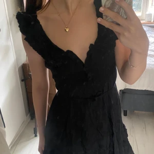 Svart volangklänning med v-ringning - Supersöt svart kortklänning med knappt fram så man kan välja ringning och volangdetaljer längs axlar och kjol. Klänningen är ärmlös och har knyt i midjan. Perfekt för dig som gillar en feminin klänning.