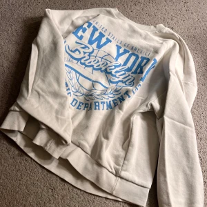 Vit sweatshirt med blå Brooklyn-tryck - Vit sweatshirt med stort blått tryck där det står 'New York Brooklyn'. Tröjan har rund hals, långa ärmar och ribbade muddar. Finns defekter på därför lågt pris