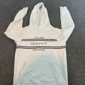 Vit hoodie från Gant med tryck - Vit hoodie från Gant med tryck 'EST. 1949', 'GANT' och 'NEW HAVEN' på bröstet. Tröjan har huva med snörning, känguruficka och marinblå samt röda ränder över bröstet. Perfekt för en chill och sportig look.
