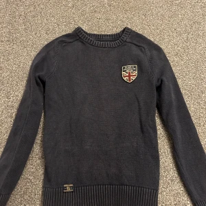 Mörkblå stickad tröja från Morris - Snygg mörkblå stickad tröja från Morris Gents i storlek XS. Tröjan har ett broderat märke med brittisk flagga på bröstet och ribbade muddar vid hals, ärmslut och nederkant. Perfekt för dig som gillar klassisk och stilren look.