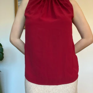 Röd ärmlös blus med knyt i nacken - Snygg röd blus med hög hals och fina rynkdetaljer framtill. Toppen är ärmlös och har ett svart sammetsband som knyts i nacken, vilket ger en elegant touch. Passformen är loose och materialet känns mjukt och följsamt.