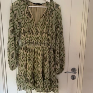 Grönmönstrad kortklänning från Zara - Supersöt kortklänning från Zara med grönt och beige mönster, v-ringning och långa ärmar. Klänningen har smock i midjan, volangdetaljer nertill och är tillverkad i ett lätt, luftigt material. 