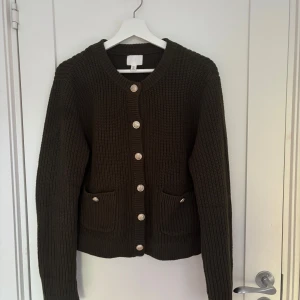 Mörkgrön stickad kofta från H&M - Snygg mörkgrön stickad kofta från H&M med grov ribbstickning, dekorativa silvriga knappar framtill och två fickor. Koftan har lång ärm och rund halsringning, perfekt för lager-på-lager under kyliga dagar.