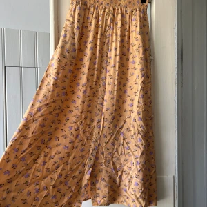 Blommig långkjol från Y.A.S i M - Superfin långkjol från Y.A.S i en ljus aprikos färg med små lila blommor. Kjolen har knappar framtill och resår i midjan för en bekväm passform. Perfekt för vår och sommar när du vill ha en luftig och söt look.