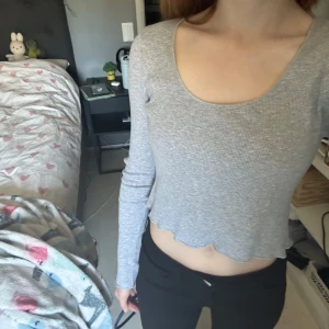 Grå croppad ribbad topp Brandy Melville - Säljer en ljusgrå croppad långärmad topp från Brandy Melville. Toppen är ribbad med rundad urringning och har vågig kant nertill för en extra söt touch. Perfekt att styla med höga jeans eller kjol.