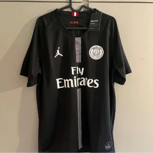 Svart PSG fotbollströja Jordan x Nike - Svart PSG fotbollströja med vita detaljer, Fly Emirates-tryck och Paris Saint-Germain-logga på bröstet. Jordan-logga på ena sidan och en smal rand i mitten. Tillverkad i lätt och ventilerande material, perfekt för träning och för sommaren.