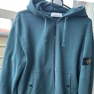 Grön hoodie innermaterial fleece från Stone Island vinter + gåva på köpet  - Snygg grön vindjacka från Stone Island med huva och ikonisk patch på ärmen. Jackan har dragkedja framtill, två fickor med knappar och en clean look som passar perfekt till streetwear. Materialet känns lätt och smidigt, perfekt för lager-på-lager. 