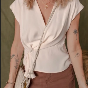 Krämvit omlottopp med knyt - Snygg krämvit omlottopp med v-ringning och knyt i sidan. Toppen har korta ärmar och är tillverkad i ett mjukt, lätt tyg som ger en avslappnad men stilren look. Perfekt att matcha med jeans eller kjol för en trendig outfit.