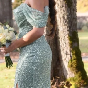 Glittrig grön långklänning offshoulder - Magisk långklänning i glittrande grönt med offshoulder-ärmar och figurnära passform. Klänningen har en snygg slits framtill och är täckt av paljetter som ger en riktigt festlig look. Perfekt för bal eller andra speciella tillfällen.